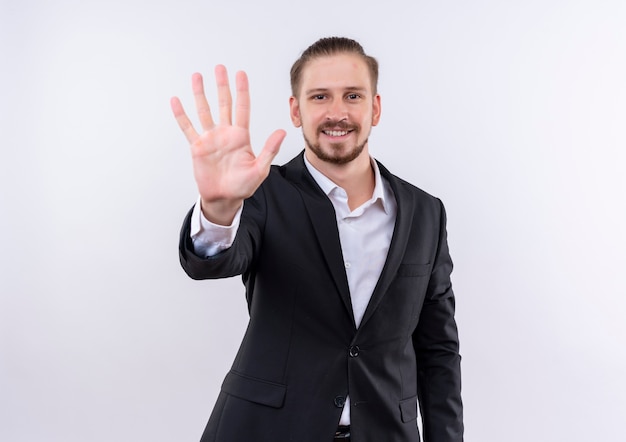 Homem de negócios bonito vestindo terno aparecendo e apontando para cima com os dedos número cinco em pé sobre um fundo branco