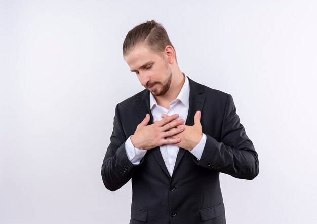 Homem de negócios bonito vestindo terno de mãos dadas no peito, sentindo emoções agradecidas e positivas com os olhos fechados, de pé sobre um fundo branco