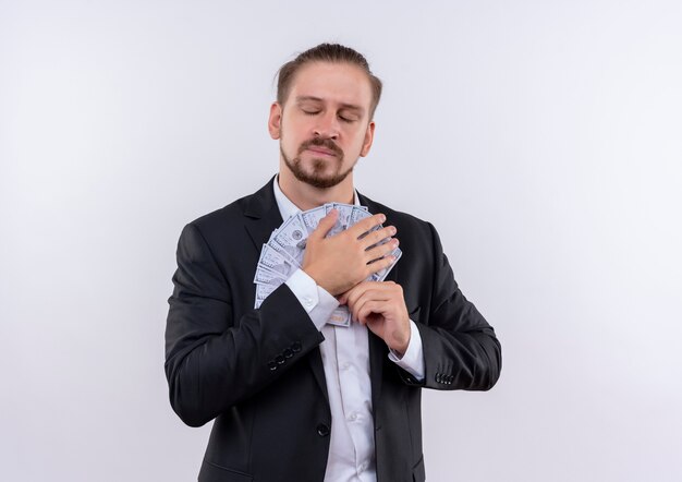 Homem de negócios bonito vestindo terno e abraçando dinheiro segurando dinheiro no peito com os olhos fechados, de pé sobre um fundo branco