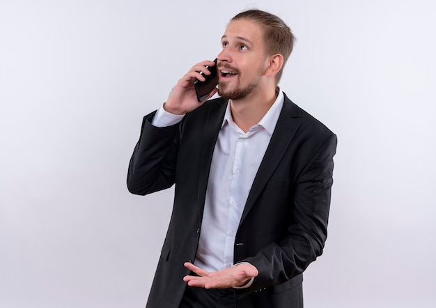 Homem de negócios bonito vestindo terno falando no celular, feliz e positivo em pé sobre um fundo branco