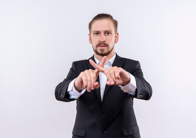 Homem de negócios bonito vestindo terno fazendo gesto de defesa cruzando os dedos indicadores com o rosto sério em pé sobre um fundo branco