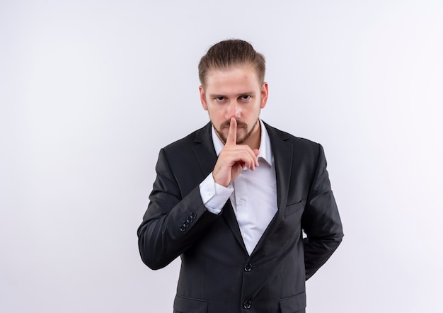 Homem de negócios bonito vestindo terno fazendo gesto de silêncio com o dedo nos lábios em pé sobre um fundo branco