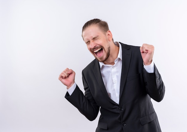 Homem de negócios bonito vestindo terno louco feliz cerrando os punhos regozijando-se com seu sucesso em pé sobre um fundo branco