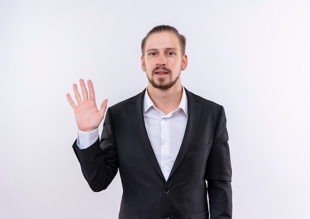 Homem de negócios bonito vestindo terno mostrando o número cinco e sorrindo confiante em pé sobre um fundo branco