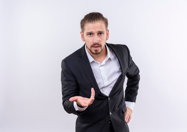 Homem de negócios bonito vestindo terno olhando para a câmera com o braço estendido enquanto faz uma pergunta em pé sobre um fundo branco