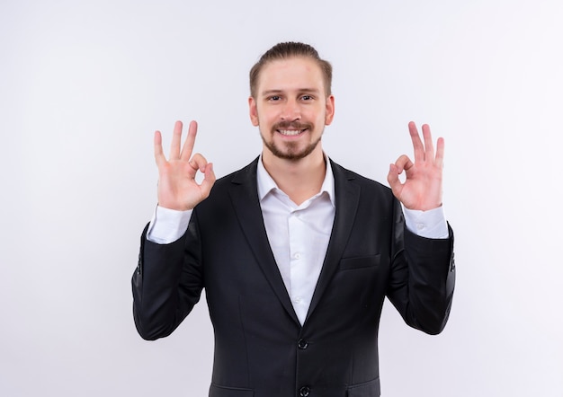 Homem de negócios bonito vestindo terno olhando para a câmera sorrindo mostrando ok cantar com as duas mãos em pé sobre um fundo branco