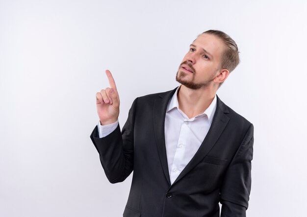 Homem de negócios bonito vestindo terno olhando para o lado com uma expressão pensativa apontando com o dedo em pé sobre um fundo branco
