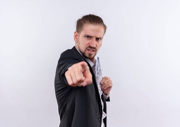 Homem de negócios bonito vestindo terno segurando dinheiro apontando com o dedo indicador para a câmera com cara de raiva em pé sobre um fundo branco