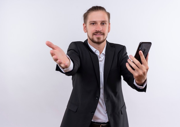 Homem de negócios bonito vestindo terno segurando smartphone fazendo gesto de boas-vindas com a mão sorrindo amigável olhando para a câmera em pé sobre fundo branco