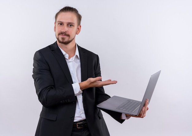 Homem de negócios bonito vestindo terno segurando um laptop apresentando-se com o braço da mão, parecendo confiante em pé sobre um fundo branco