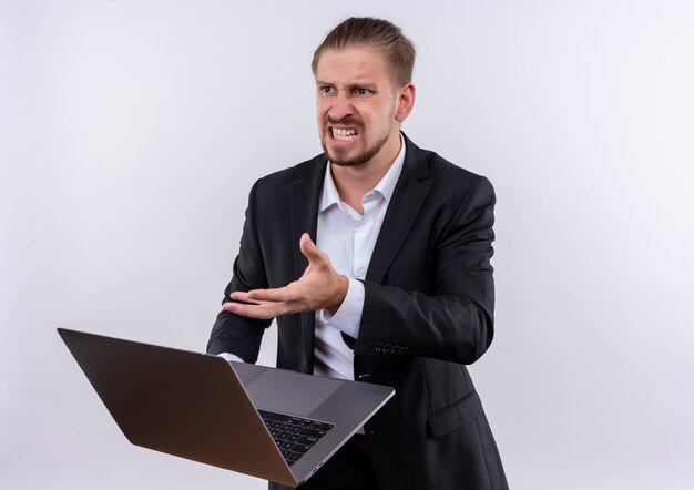 Homem de negócios bonito vestindo terno segurando um laptop e parecendo frustrado com uma cara de raiva em pé sobre um fundo branco