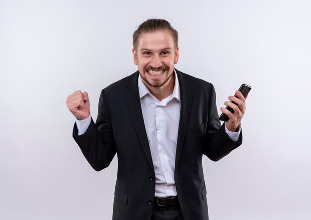 Homem de negócios bonito vestindo terno segurando um smartphone com o punho cerrado, feliz e animado em pé sobre um fundo branco