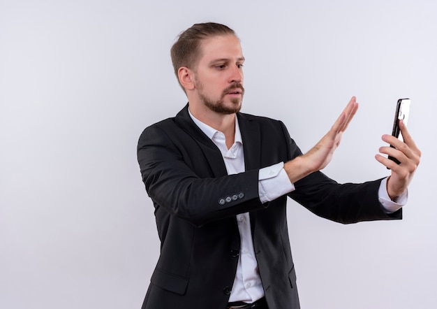 Homem de negócios bonito vestindo terno segurando um smartphone olhando para a tela fazendo um gesto de calma com o braço em pé sobre um fundo branco