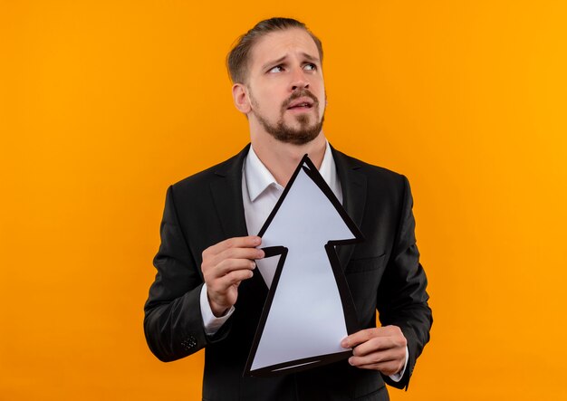 Foto grátis homem de negócios bonito vestindo terno segurando uma seta branca, olhando para cima confuso em pé sobre um fundo laranja