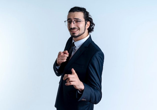 Homem de negócios de terno preto e óculos apontando com o dedo indicador para a frente sorrindo alegremente em pé sobre uma parede branca