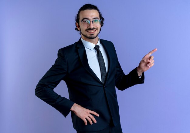 Homem de negócios de terno preto e óculos apontando com o dedo indicador para o lado sorrindo alegremente em pé sobre a parede azul