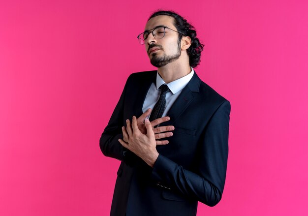 Homem de negócios de terno preto e óculos com as mãos cruzadas sobre o peito olhando para o lado e sentindo-se grato em pé sobre a parede rosa