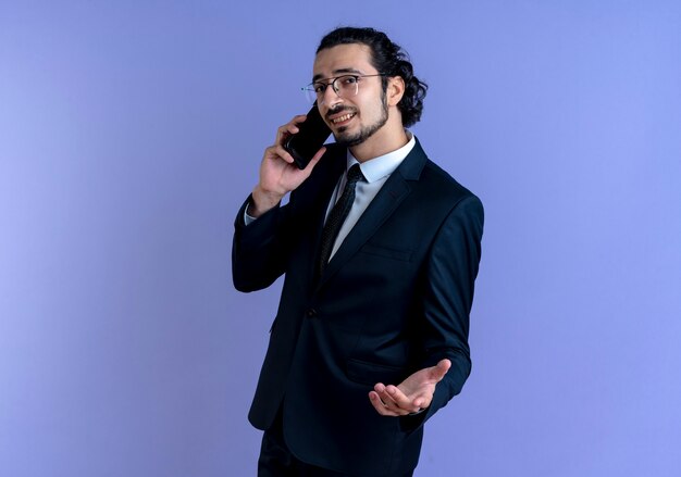 Homem de negócios de terno preto e óculos falando no celular parecendo descontente em pé sobre a parede azul