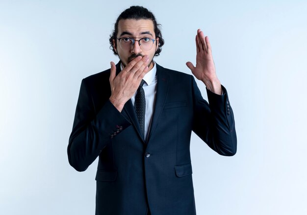Homem de negócios de terno preto e óculos levantando a mão parecendo surpreso cobrindo a boca com a mão em pé sobre a parede branca