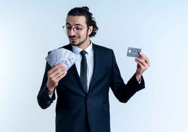 Homem de negócios de terno preto e óculos, mostrando dinheiro e cartão de crédito, sorrindo com uma cara feliz em pé sobre uma parede branca