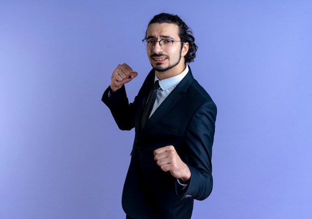 Homem de negócios de terno preto e óculos olhando para a frente com o punho cerrado, posando como um boxeador em pé sobre uma parede azul