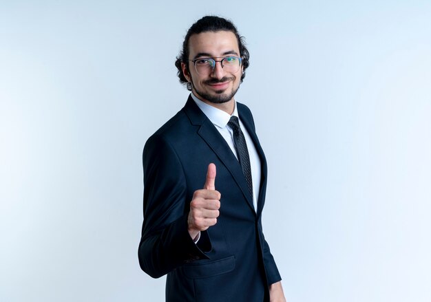 Homem de negócios de terno preto e óculos olhando para a frente com uma expressão confiante sorrindo mostrando os polegares em pé sobre uma parede branca
