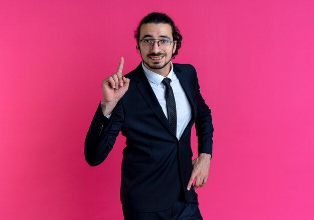 Homem de negócios de terno preto e óculos olhando para a frente, mostrando o dedo indicador sorrindo, tendo uma ótima ideia em pé sobre a parede rosa