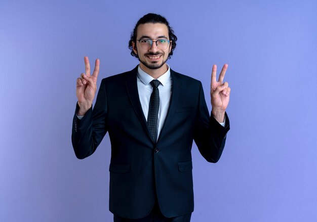 Homem de negócios de terno preto e óculos olhando para a frente sorrindo alegremente, mostrando o sinal da vitória com as duas mãos em pé sobre a parede azul