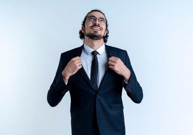 Homem de negócios de terno preto e óculos olhando para cima satisfeito consertando seu terno em pé sobre uma parede branca