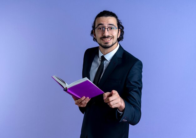 Homem de negócios de terno preto e óculos segurando um caderno apontando com o dedo indicador para a frente, sorrindo alegremente em pé sobre a parede azul