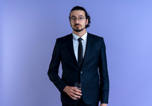 Homem de negócios de terno preto e óculos segurando um copo d'água olhando para a frente com uma cara séria em pé sobre a parede azul
