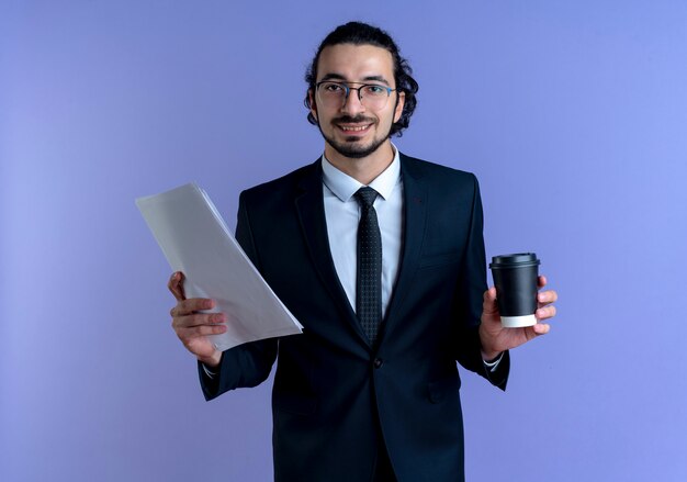 Homem de negócios de terno preto e óculos segurando uma xícara de café e documentos olhando para a frente sorrindo alegremente em pé sobre a parede azul