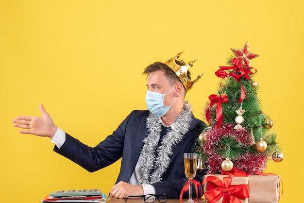 Homem de negócios de vista frontal dando a mão sentado à mesa perto da árvore de natal e presentes em fundo amarelo