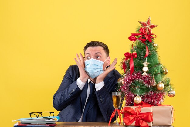 Homem de negócios espantado de vista frontal sentado à mesa perto da árvore de natal e presentes no espaço livre de fundo amarelo