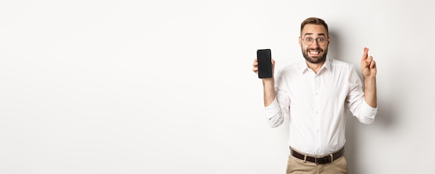 Homem de negócios jovem esperançoso mostrando a tela do celular segurando os dedos cruzados esperando por resultados online