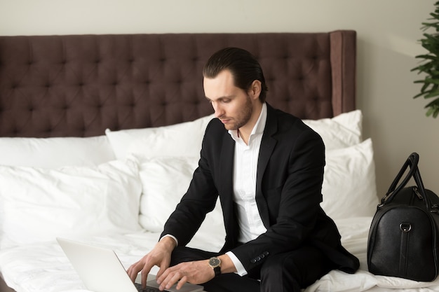 Homem de negócios novo que senta-se na cama que trabalha no portátil no hotel.