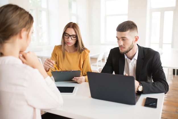 Homem de negócios pensativo com laptop e mulher de negócios em óculos com pasta na mão falando com candidato sobre trabalho jovens empregadores pensativos passando entrevista de emprego no escritório moderno