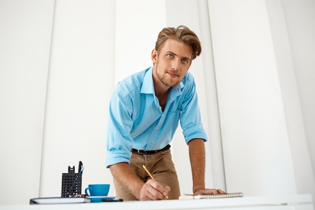 Homem de negócios pensativo seguro considerável novo que está de pé na tabela sobre a vista do bloco de notas. Interior de escritório moderno branco.