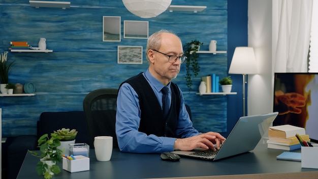 Homem de negócios sênior remoto abrindo laptop e lendo relatórios trabalhando em casa tomando café