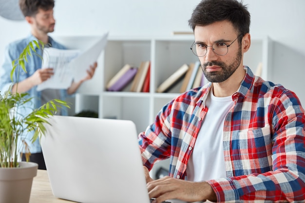 Homem de negócios sério com barba espessa, analisa tabelas de renda e gráficos em computador laptop, vestido com camisa xadrez