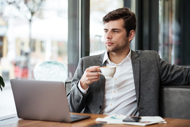 Homem de negócios sério sentado junto à mesa no café com o computador portátil enquanto bebe café e olhando para a janela