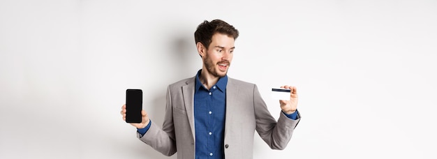 Foto grátis homem de negócios sorridente de compras on-line de terno mostrando cartão de crédito de plástico com tela de smartphone vazia