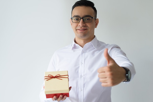 Homem de negócios sorridente segurando a caixa de presente e mostrando o polegar para cima