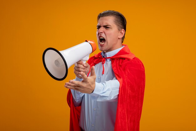 Homem de negócios super-herói com capa vermelha gritando para o megafone com expressão agressiva e braço estendido sobre a parede laranja