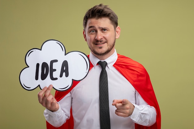 Homem de negócios super-herói com capa vermelha segurando um cartaz de bolha do discurso com a ideia da palavra apontando com o dedo indicador para a câmera, sorrindo em pé sobre um fundo verde