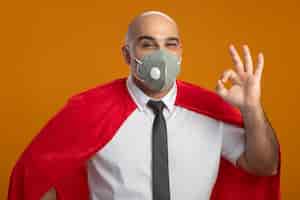 Foto grátis homem de negócios super-herói com máscara facial protetora e capa vermelha olhando para frente sorrindo e mostrando uma placa de ok em pé sobre a parede laranja