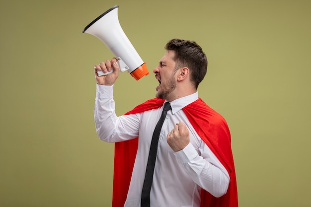 Homem de negócios super-herói furioso com capa vermelha gritando para o megafone com o punho cerrado agressivo de pé sobre um fundo verde