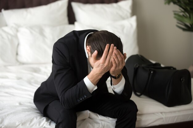 Homem de negócios virado forçado que senta-se na cama, tendo a dor de cabeça.