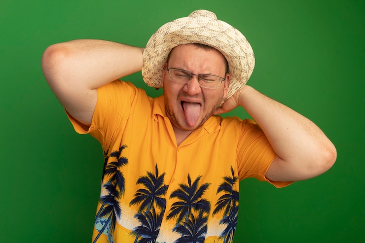 Foto grátis homem de óculos com camisa laranja e chapéu de verão mostrando a língua com expressão irritada em pé sobre a parede verde