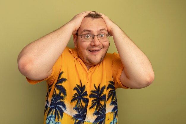 Homem de óculos com camisa laranja feliz e animado com as mãos na cabeça em pé sobre a parede verde